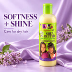 A bottle of Kids Original Naturals Shea Butter Moisturizing Hair Lotion stands on a lavender satin surface with lilac flowers. The bottle is pale yellow with a bright green cap and a lime green label featuring the product name and a photo of two children.