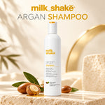 A white bottle of milk shake Argan Shampoo stands on a stone surface with argan nuts, leaves, and golden oil props, against a warm beige background with a blurred gold object. Large text at the top reads 'milk shake ARGAN SHAMPOO'.