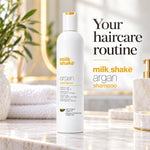 A bottle of Milk Shake Argan Shampoo stands on a marble bathroom counter with towels, a plant, and gold fixtures in the background. Large text on the right reads, 'Your haircare routine milk shake argan shampoo.