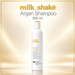 A 300 ml bottle of milk shake Argan Shampoo centered on a warm beige gradient background with a thin gold border. The bottle is white with orange and black text, and the brand name 'milk shake' is displayed prominently at the top.